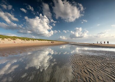 Der Himmel spiegelt sich in der letzten Pfuetze die die Ebbe uebrig gelassen hat Menschen spazieren ueber den Strand | © Gettyimages.com/olharohulya