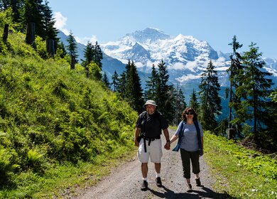 Reifes Paar, das Haendchen haelt, wandert einen Bergpfad hinunter mit schneebedeckten Bergen hinter sich. Im Berner Oberland bei Wengen, Schweiz. | © Gettyimages.com/OGphoto