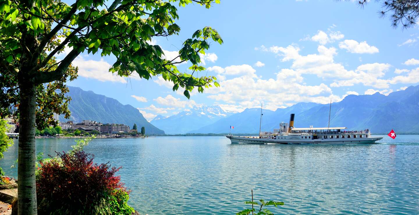 Blick von Montreux auf den Genfersee mit vorbeifahrendem Ausflugsschiff bei Sonnenschein und leichten Wolken | © Gettyimages.com/alxpin
