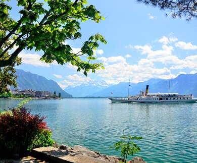 Blick von Montreux auf den Genfersee mit vorbeifahrendem Ausflugsschiff bei Sonnenschein und leichten Wolken | © Gettyimages.com/alxpin