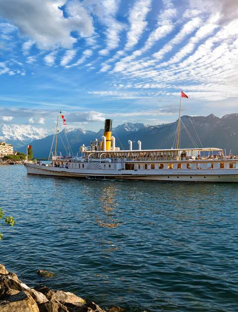 Wunderschoene Sommerabendlandschaft des Genfersees mit malerischen Ufern und Ausflugsschiff gegen alpine Berge in den Strahlen der untergehenden Sonne in Montreux, Schweiz | © Gettyimages.com/connect11