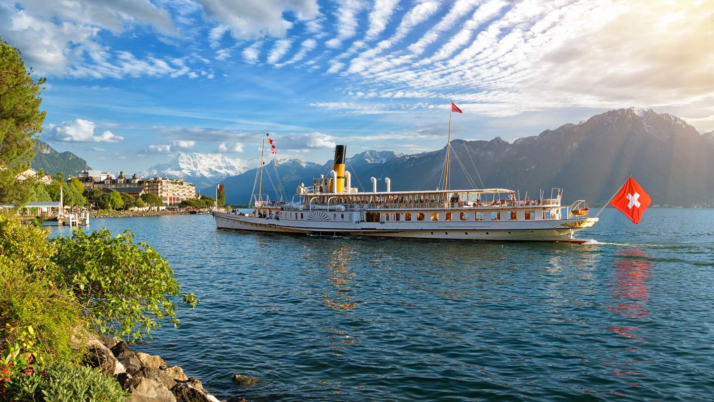 Wunderschoene Sommerabendlandschaft des Genfersees mit malerischen Ufern und Ausflugsschiff gegen alpine Berge in den Strahlen der untergehenden Sonne in Montreux, Schweiz | © Gettyimages.com/connect11