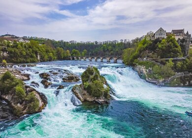 Luftaufnahme der reissenden Fluten des Rheinfalls in der Gemeinde Neuhausen am Rheinfall im Schweizer Kanton Schaffhausen mit vielen Besucher:innen auf den Aussichtsplattformen und einem Aussichtsboot | © Gettyimages.com/Arkadij Schell