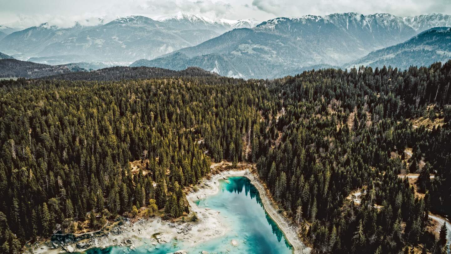 Luftaufnahme des türkisblauen Caumasees in Flims umgeben von Tannen und im Hintergrund die Alpengigipfel. | © Schweiz Tourismus/Silvan Widmer