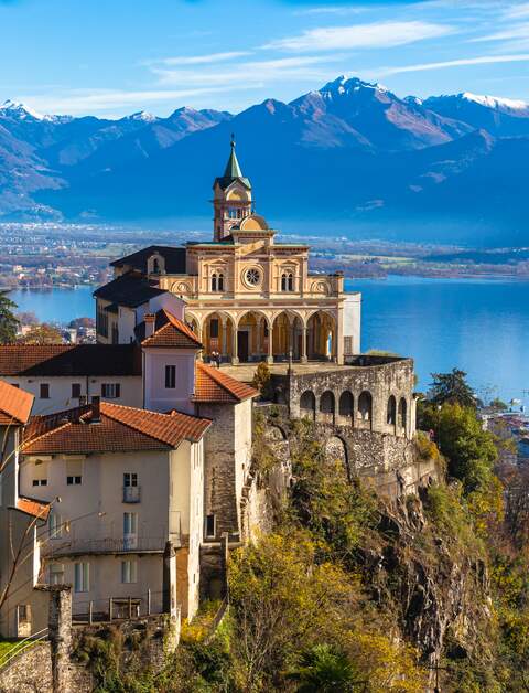 Atemberaubende Aussicht auf die Kirche Madonna del Sasso oberhalb der Stadt Locarno mit Lago Maggiore, schneebedecktem Schweizer Alpenberg und blauer Himmelswolke im Hintergrund im Herbst in der Schweiz | © Gettyimages.com/vogelisp