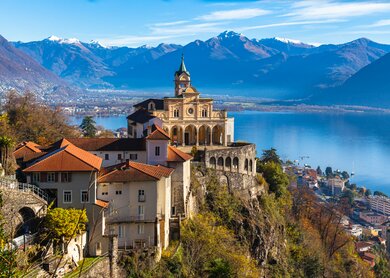 Atemberaubende Aussicht auf die Kirche Madonna del Sasso oberhalb der Stadt Locarno mit Lago Maggiore, schneebedecktem Schweizer Alpenberg und blauer Himmelswolke im Hintergrund im Herbst in der Schweiz | © Gettyimages.com/vogelisp
