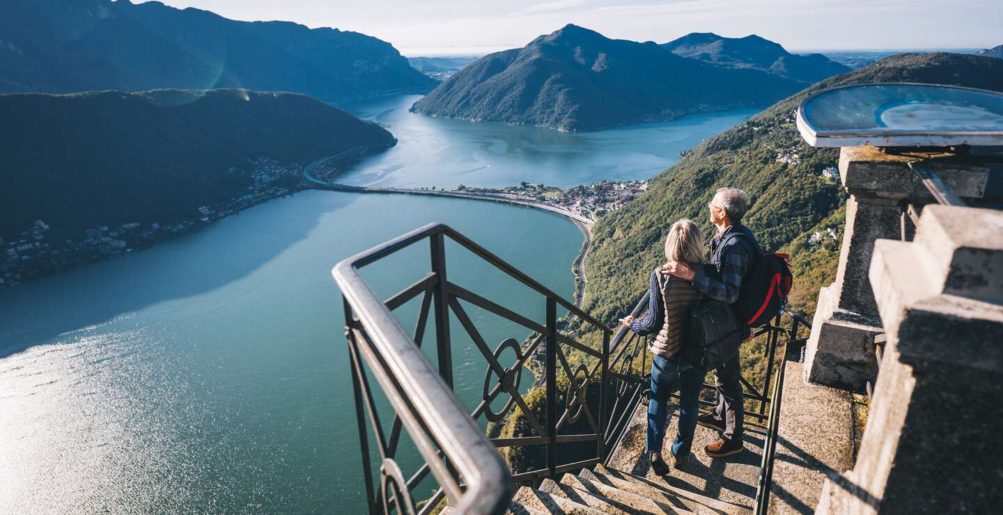 Ein reifes Paar wandert morgens über den Luganersee, haelt auf Stufen inne und blickt auf die entfernte Szene. | © Gettyimages.com/AscentXmedia