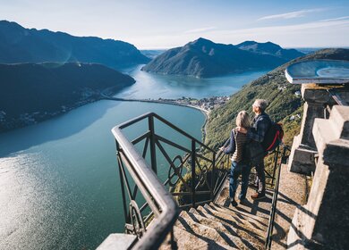 Ein reifes Paar wandert morgens über den Luganersee, haelt auf Stufen inne und blickt auf die entfernte Szene. | © Gettyimages.com/AscentXmedia