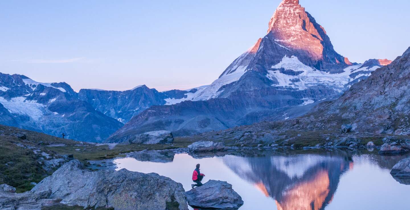 Fruehmorgens Sonnenaufgangsszene auf dem Matterhorn, das sich rosa im See spiegelt, mit Mann auf einem Felsen mit roter Daunenjacke und klarem blauen Himmel  | © Gettyimages.com/wilpunt