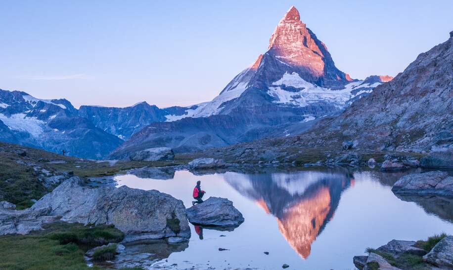 Fruehmorgens Sonnenaufgangsszene auf dem Matterhorn, das sich rosa im See spiegelt, mit Mann auf einem Felsen mit roter Daunenjacke und klarem blauen Himmel  | © Gettyimages.com/wilpunt