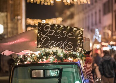 Straßenfahrzeug mit Weihnachtsdekoration und ‚Gasse Drinks‘-Schild in nächtlicher Fußgängerzone. | © Elias Bötticher