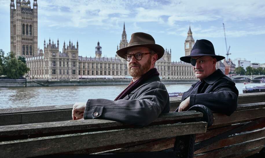 Zwei Männer, unter anderem Gabriel Book, sitzen auf einer Bank vor Westminster Abbey in der Serie "Bookish" | © polyband/Beta Film/UK TV