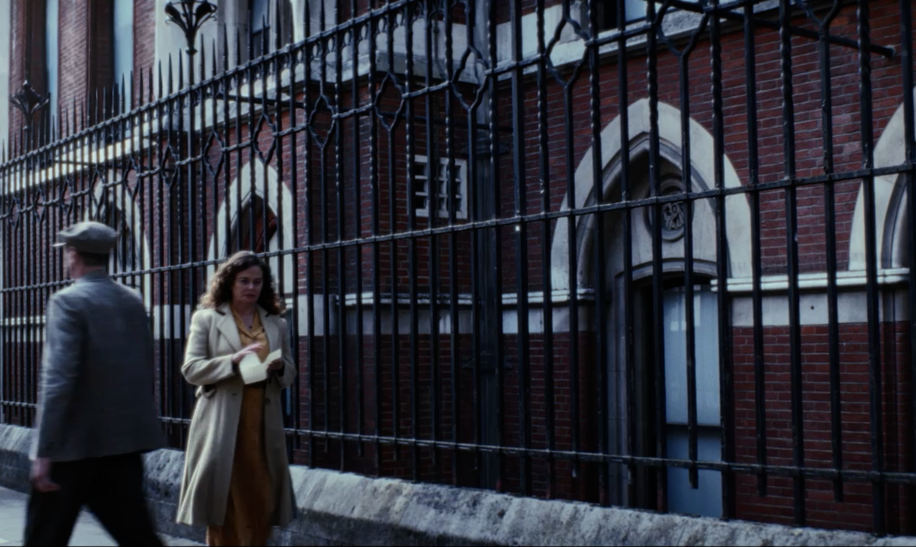 Eine Szene vor einem großen Backsteingebäude mit hohen, schmiedeeisernen Zaunelementen im Vordergrund. Die Fassade zeigt mehrere schmale, bogenförmige Fenster mit hellem Steinrahmen. Auf dem Gehweg gehen mehrere Personen aneinander vorbei, alle in historischer Straßenkleidung. Die Umgebung wirkt wie ein städtisches Viertel mit älteren, repräsentativen Gebäuden. | © polyband/Beta Film/UK TV