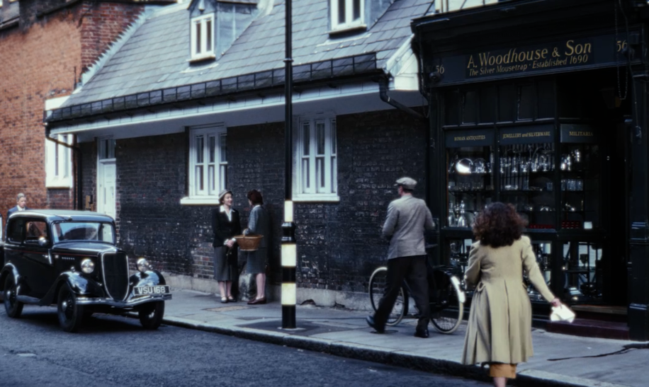 Eine Straßenszene im historischen Stil: Entlang einer schmalen Kopfsteinpflasterstraße stehen mehrere ältere Autos geparkt. Auf dem Gehweg unterhalten sich mehrere Personen in zeittypischer Kleidung. Rechts befindet sich ein traditionelles Geschäft mit dunkler Fassade und goldfarbener Beschriftung, auf der „A. Woodhouse & Son — The Silver Showroom Established 1900“ zu lesen ist. Eine Person nähert sich von hinten dem Laden, während eine andere Person mit Fahrrad vorbeigeht. Die Gebäude bestehen überwiegend aus Backstein und haben weiße Fensterrahmen. | © polyband/Beta Film/UK TV