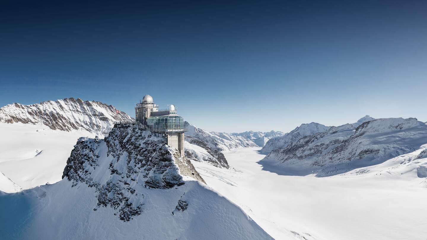 Hochgelegenes Gebäude auf einem verschneiten Berggipfel mit Kuppelbau. Umgeben von schneebedeckten, zerklüfteten Berglandschaften unter klarem, blauem Himmel. | © Jungfraubahnen