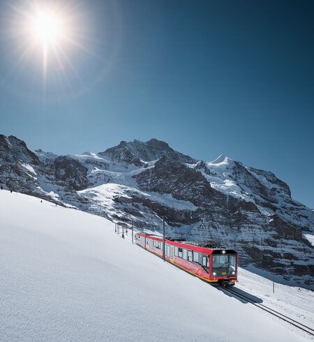 Ein roter Zug fährt eine verschneite Berghangstrecke entlang, umgeben von majestätischen, schneebedeckten Gipfeln unter klarem, blauem Himmel. Die Sonne scheint hell und taucht die Winterlandschaft in warmes Licht. | © Jungfraubahnen, Jeroen Seyffer Fotografie
