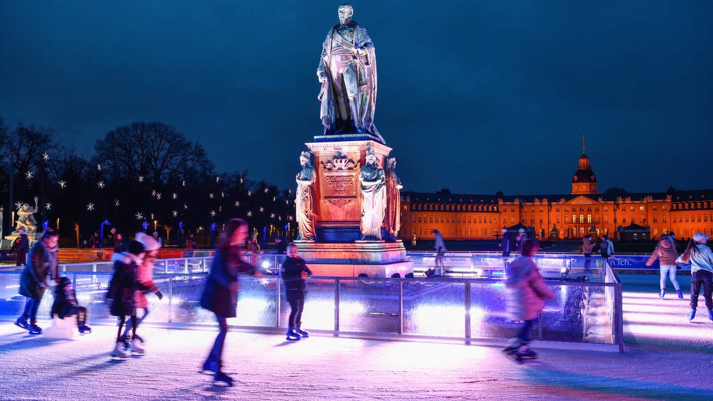 Große Statue einer historischen Figur auf einem kunstvoll verzierten Sockel inmitten einer beleuchteten Eislaufbahn bei Nacht, umgeben von Schlittschuhläufern und einem eindrucksvoll beleuchteten Gebäude im Hintergrund. | © KTG Karlsruhe Tourismus GmbH, Fabian von Poser