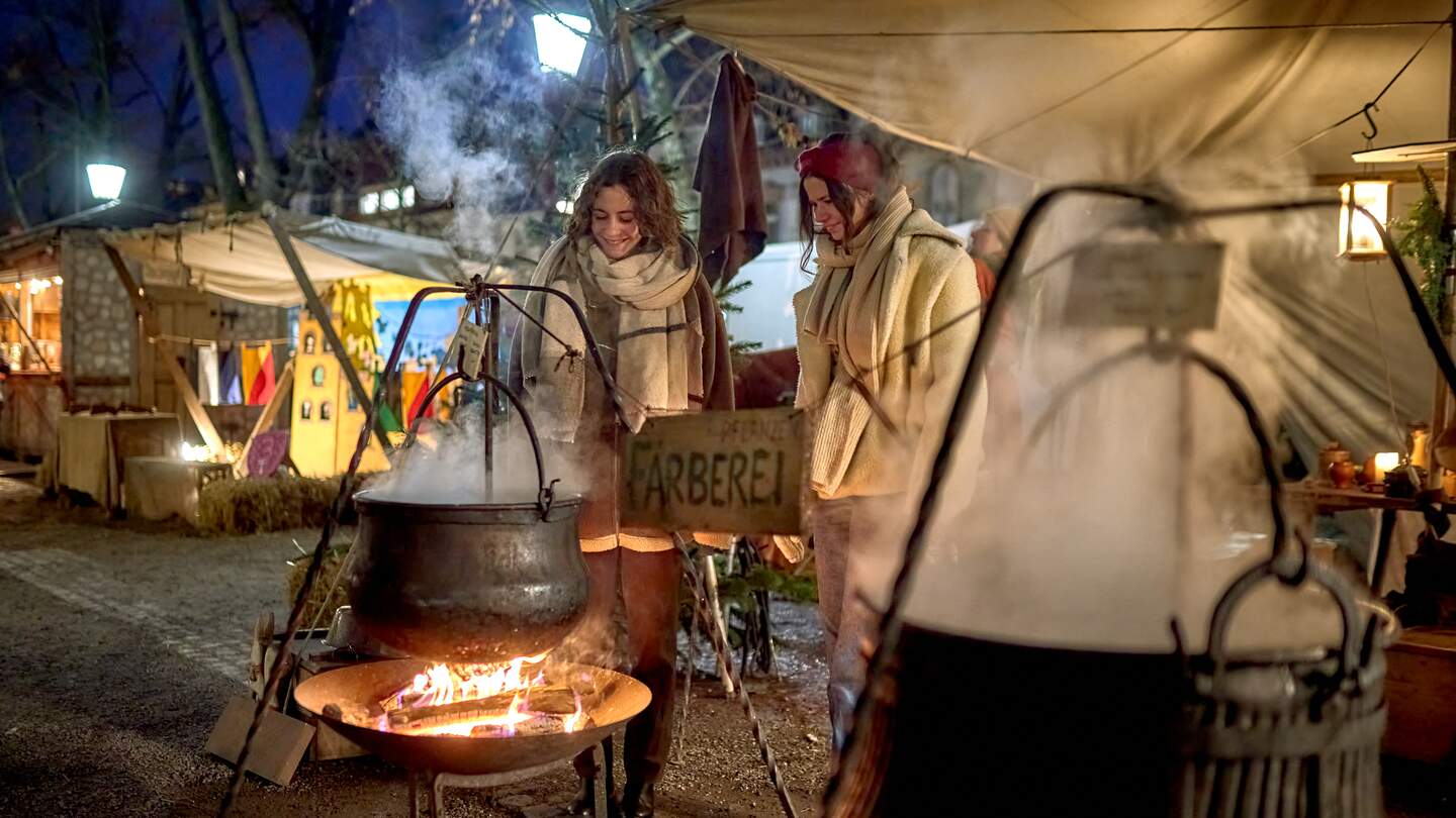 Zwei Personen stehen neben einem großen schwarzen Kessel, der über offenem Feuer hängt und aus dem Dampf aufsteigt. Die Szene spielt sich auf einem abendlichen, mittelalterlichen Weihnachtsmarkt ab, mit beleuchteten Ständen im Hintergrund. | © KTG Karlsruhe Tourismus GmbH, Bruno Kelzer
