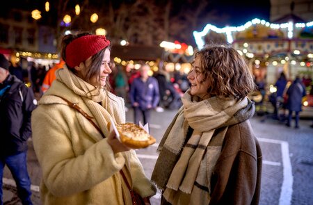 Zwei Personen stehen auf einem festlich beleuchteten Markt im Freien. Eine Person hält einen Teller mit einem Lebensmittel und bietet es der anderen an. Beide sind warm gekleidet, was auf kaltes Wetter hinweist. Im Hintergrund sind Lichter, Dekorationen und weitere Besucher zu sehen. | © KTG Karlsruhe Tourismus GmbH, Bruno Kelzer