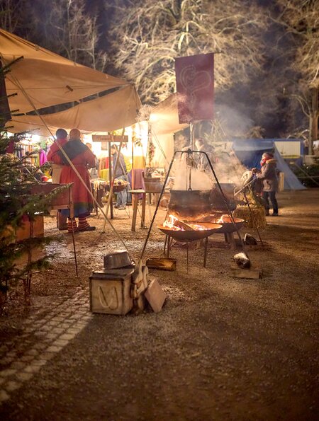 Abendliche Szene auf einem mittelalterlichen Weihnachtsmarkt mit offenen Feuerstellen, einem großen Kessel, Marktständen unter Zelten und Personen in historischer Kleidung. Die warme Beleuchtung und winterliche Kulisse mit kahlen Bäumen schaffen eine festliche und authentische Atmosphäre. | © KTG Karlsruhe Tourismus GmbH, Bruno Kelzer