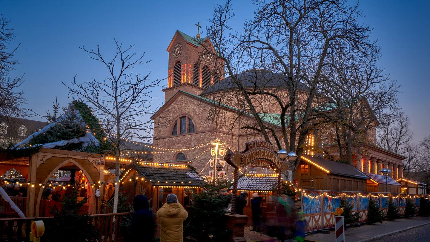 Festlich beleuchteter Weihnachtsmarkt vor einer großen Kirche mit Uhrturm und Kreuz, umgeben von Holzständen, Lichterketten und winterlich kahlen Bäumen. Besucher schlendern durch die stimmungsvolle Abendkulisse. | © KTG Karlsruhe Tourismus GmbH, Bruno Kelzer