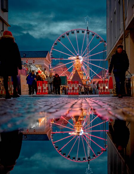 Straßenszene bei Dämmerung mit einem farbig beleuchteten Riesenrad im Zentrum, dessen Spiegelung in einer Pfütze auf dem Kopfsteinpflaster sichtbar ist. Menschen flanieren entlang der Straße, umgeben von beleuchteten Gebäuden und dramatisch bewölktem Himmel. | © KTG Karlsruhe Tourismus GmbH, Bruno Kelzer