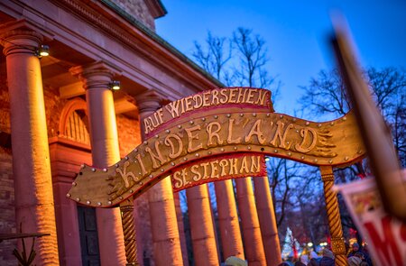 Beleuchteter Torbogen mit der Aufschrift ‚AUF WIEDERSEHEN KINDERLAND ST. STEPHAN‘ vor einem Gebäude mit Säulen und Bäumen, aufgenommen bei abendlicher Stimmung. | © KTG Karlsruhe Tourismus GmbH, Bruno Kelzer