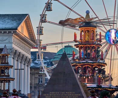 Festliche Szene eines Weihnachtsmarkts mit beleuchtetem Riesenrad, kunstvoll dekorierter Weihnachtspyramide, klassischem Gebäude mit Säulen und Schneebedeckung, sowie einem leuchtenden Kugelobjekt und einem Denkmal im Vordergrund. | © KTG Karlsruhe Tourismus GmbH, Bruno Kelzer