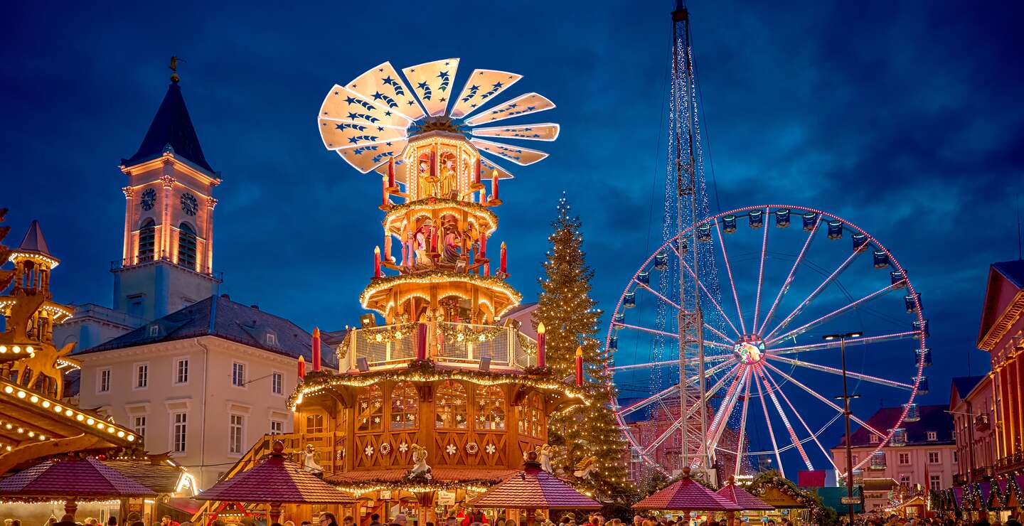 Festliche Szene auf einem Weihnachtsmarkt mit einer großen, beleuchteten Weihnachtspyramide, einem hell erleuchteten Riesenrad im Hintergrund und einem Kirchturm mit Uhr. Zahlreiche Besucher schlendern durch die abendliche Marktstimmung. | © KTG Karlsruhe Tourismus GmbH, Bruno Kelzer