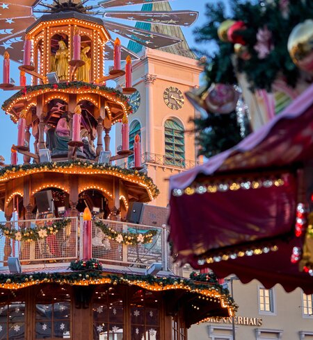 Weihnachtsmarkt mit großer, mehrstöckiger, beleuchteter Weihnachtspyramide mit Figuren und rotierenden Flügeln, im Hintergrund eine Kirche mit Uhrturm und im Vordergrund ein dekoriertes Element mit Girlanden und Ornamenten. | © KTG Karlsruhe Tourismus GmbH, Bruno Kelzer