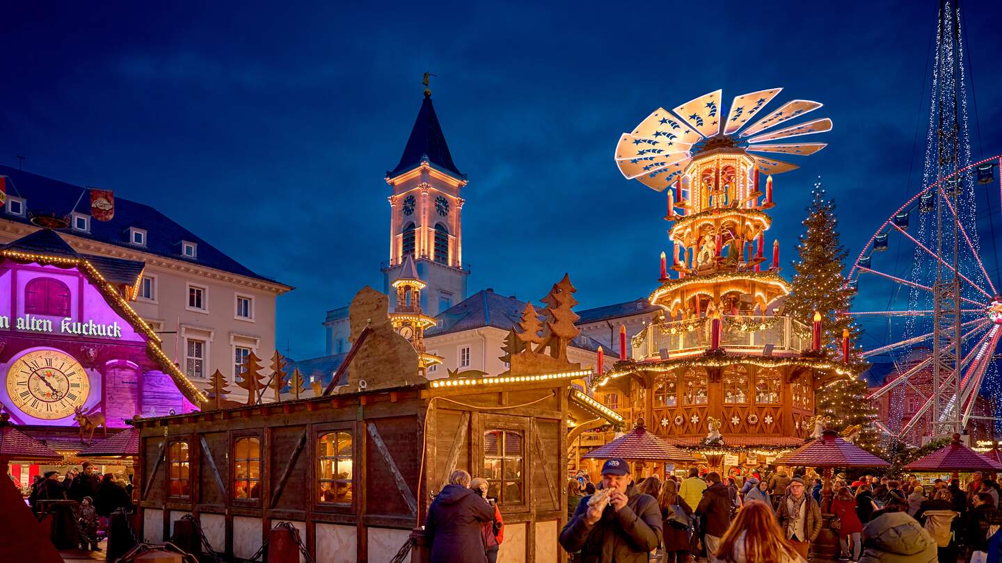 Festliche Szene auf einem Weihnachtsmarkt mit einer großen, beleuchteten Weihnachtspyramide, einem hell erleuchteten Riesenrad im Hintergrund und einem Kirchturm mit Uhr. Zahlreiche Besucher schlendern durch die abendliche Marktstimmung. | © KTG Karlsruhe Tourismus GmbH, Bruno Kelzer