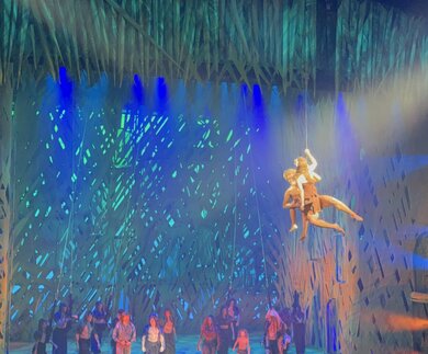 Blick auf eine große Theaterbühne mit farbigem Lichtspiel in Grün‑ und Blautönen. Im Mittelpunkt schweben zwei Artistinnen oder Artisten an Seilen in der Luft, während unten ein Ensemble in dunklen Kostümen auf der Bühne steht. Im Vordergrund sitzt das Publikum und beobachtet die akrobatische Szene. | © Michelle Khan