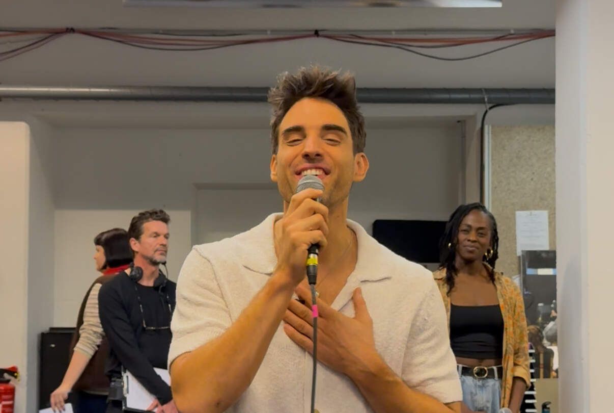 Eine Person steht im Probenraum und singt in ein Mikrofon, während eine Hand auf der Brust liegt. Im Hintergrund stehen weitere Personen, ebenfalls in den Probenprozess eingebunden. Der Raum ist mit technischen Geräten und Kabeln ausgestattet und vermittelt eine typische Studio‑ oder Probensituation. | © Michelle Khan