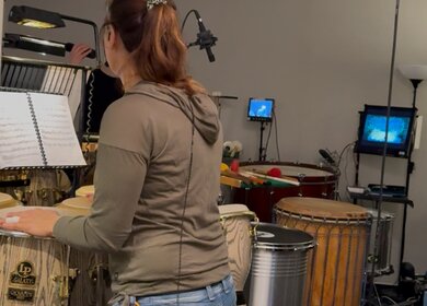 Eine Person steht in einem Probenraum zwischen mehreren großen Percussion‑Instrumenten wie Congas, Trommeln und Metalltrommeln. Vor ihr liegt ein aufgeschlagenes Notenheft auf einem Ständer. Im Hintergrund sind weitere Instrumente sowie kleine Monitore sichtbar, die eine Bühnen- oder Videoansicht zeigen. | © Michelle Khan