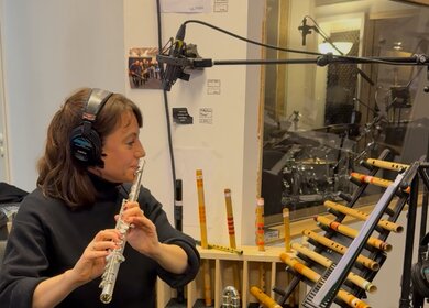 Eine Person sitzt in einem Probenraum und spielt eine Querflöte. Sie trägt große Kopfhörer und sitzt vor mehreren Mikrofonen sowie einem Notenständer mit Tablet. Hinter ihr stehen verschiedene Holzblasinstrumente in Halterungen, darunter Flöten und Pfeifen in unterschiedlichen Größen. Im Hintergrund ist durch eine Glasscheibe ein weiterer Raum mit einem Schlagzeug zu sehen. | © Michelle Khan