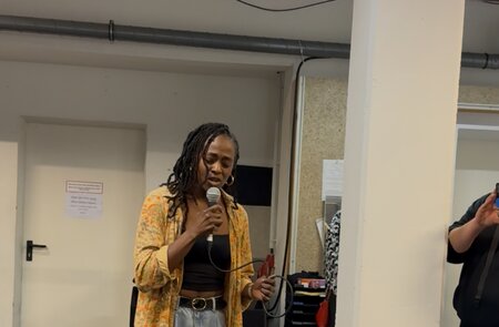 Eine Person steht in einem Probenraum und singt in ein Mikrofon. Sie trägt eine weit geschnittene Jeans, ein schwarzes Top und ein gelb gemustertes Hemd. In der Hand hält sie das Mikrofonkabel.  | © Michelle Khan