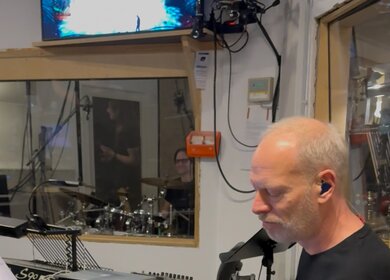 Eine Person sitzt in einem Probenraum und spielt eine E‑Gitarre. Vor ihr stehen Notenständer, Mischpultgeräte und verschiedene technische Instrumente. Hinter einer Glasscheibe ist ein separater Raum mit einem Schlagzeug zu sehen. Kabel, Mikrofone und Bildschirme gehören zur professionellen Studio‑ oder Probenumgebung. | © Michelle Khan