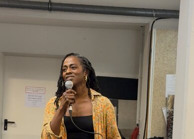 Eine Person steht in einem Probenraum und singt in ein Mikrofon. Sie trägt eine weit geschnittene Jeans, ein schwarzes Top und ein gelb gemustertes Hemd. In der Hand hält sie das Mikrofonkabel.  | © Michelle Khan