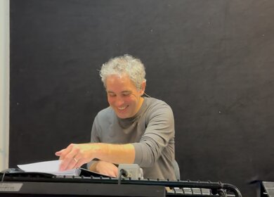 Eine Person sitzt an einem Tasteninstrument und hält mit einer Hand ein Blatt Papier, das vor ihr liegt. Der Hintergrund ist schlicht und dunkel, wodurch der Fokus auf der Tätigkeit der Person am Instrument liegt. | © Michelle Khan