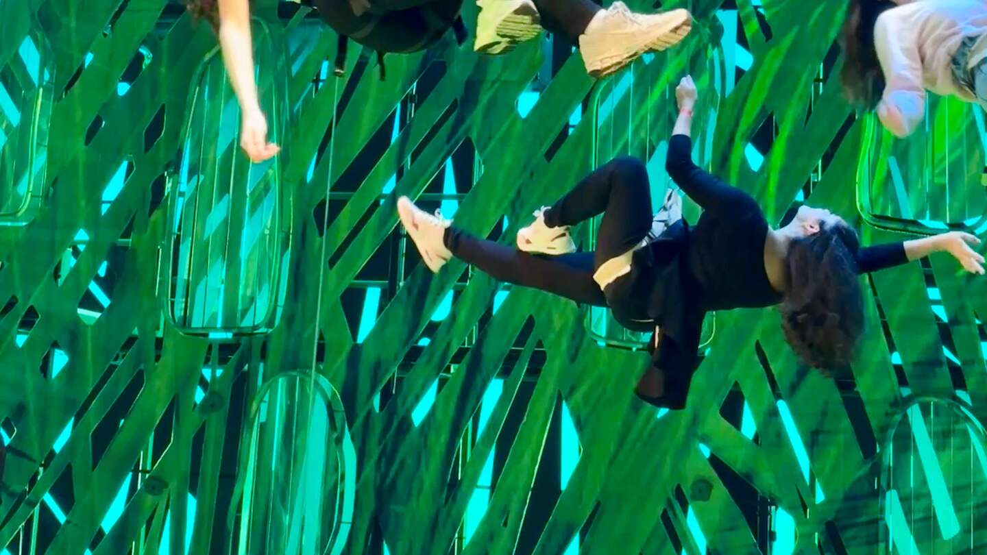 Menschen hängen in akrobatischen Positionen an Seilen oder Gurten vor einer Bühne mit intensiv grün-blau beleuchtetem Hintergrund. Eine der Personen schwebt seitlich gedreht in der Luft, mit ausgestrecktem Arm und angewinkelten Beinen, während andere Körper oberhalb im Bildrand sichtbar sind. | © Privat