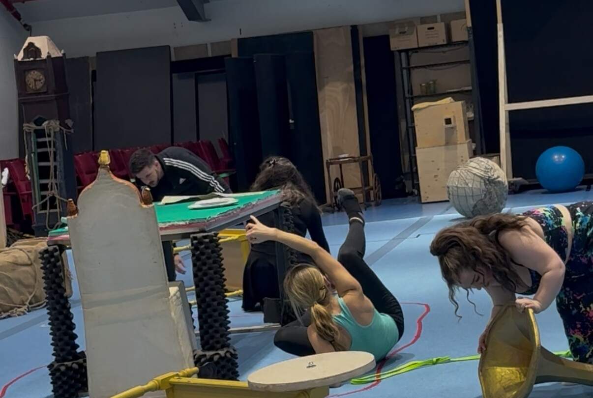Mehrere Personen probieren in einem Probenraum szenische Bewegungen aus. Sie liegen, knien oder beugen sich in verschiedene Richtungen, während Requisiten wie ein gelber Kasten, ein großer Ball, Stoffelemente und eine bemalte Spielfläche im Raum verteilt sind.  | © Noelle Löw