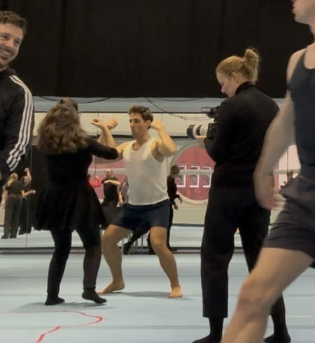 Mehrere Personen befinden sich in einem Probenraum. Eine Person in Sportkleidung übt eine dynamische Pose oder Bewegung, während eine andere davor steht und Anweisungen gibt. | © Michelle Khan