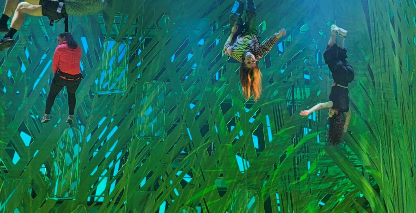 Mehrere Personen hängen gesichert an Seilen und schweben in unterschiedlichen akrobatischen Positionen vor einer großflächigen, hell beleuchteten grünen Bühnenkulisse. Einige sind kopfüber oder seitlich gedreht, während andere sich in der Luft stabilisieren. Unten am Bühnenrand steht eine weitere Person und beobachtet die Szene. Theatralisches Licht und die abstrakte, grasähnliche Struktur des Hintergrunds erzeugen eine dynamische, schwebende Atmosphäre. | © Michelle Khan