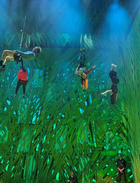 Mehrere Personen hängen gesichert an Seilen und schweben in unterschiedlichen akrobatischen Positionen vor einer großflächigen, hell beleuchteten grünen Bühnenkulisse. Einige sind kopfüber oder seitlich gedreht, während andere sich in der Luft stabilisieren. Unten am Bühnenrand steht eine weitere Person und beobachtet die Szene. Theatralisches Licht und die abstrakte, grasähnliche Struktur des Hintergrunds erzeugen eine dynamische, schwebende Atmosphäre. | © Michelle Khan