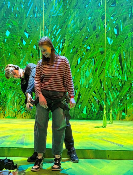 Eine Person steht auf einer hell beleuchteten Bühne mit grün‑blauer, abstrakter Kulisse. Sie trägt ein gestreiftes Oberteil, Jeans und Sportschuhe und wird von einer zweiten Person dabei unterstützt, ein Sicherungsharness anzulegen. Auf dem Boden liegen weitere Gurte und Ausrüstungsteile bereit. | © Michelle Khan