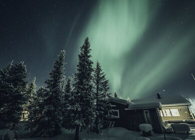 Nordlichter über einer schneebedeckten Winterlandschaft mit einem Holzhaus im Hintergrund. | © imagebank.sweden.se/Asaf Kliger