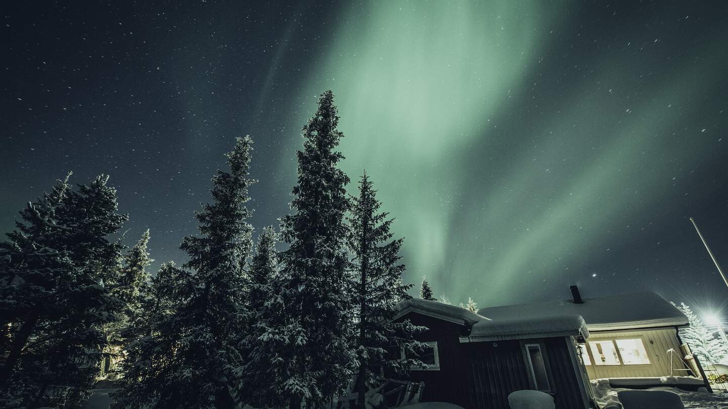 Nordlichter über einer schneebedeckten Winterlandschaft mit einem Holzhaus im Hintergrund. | © imagebank.sweden.se/Asaf Kliger