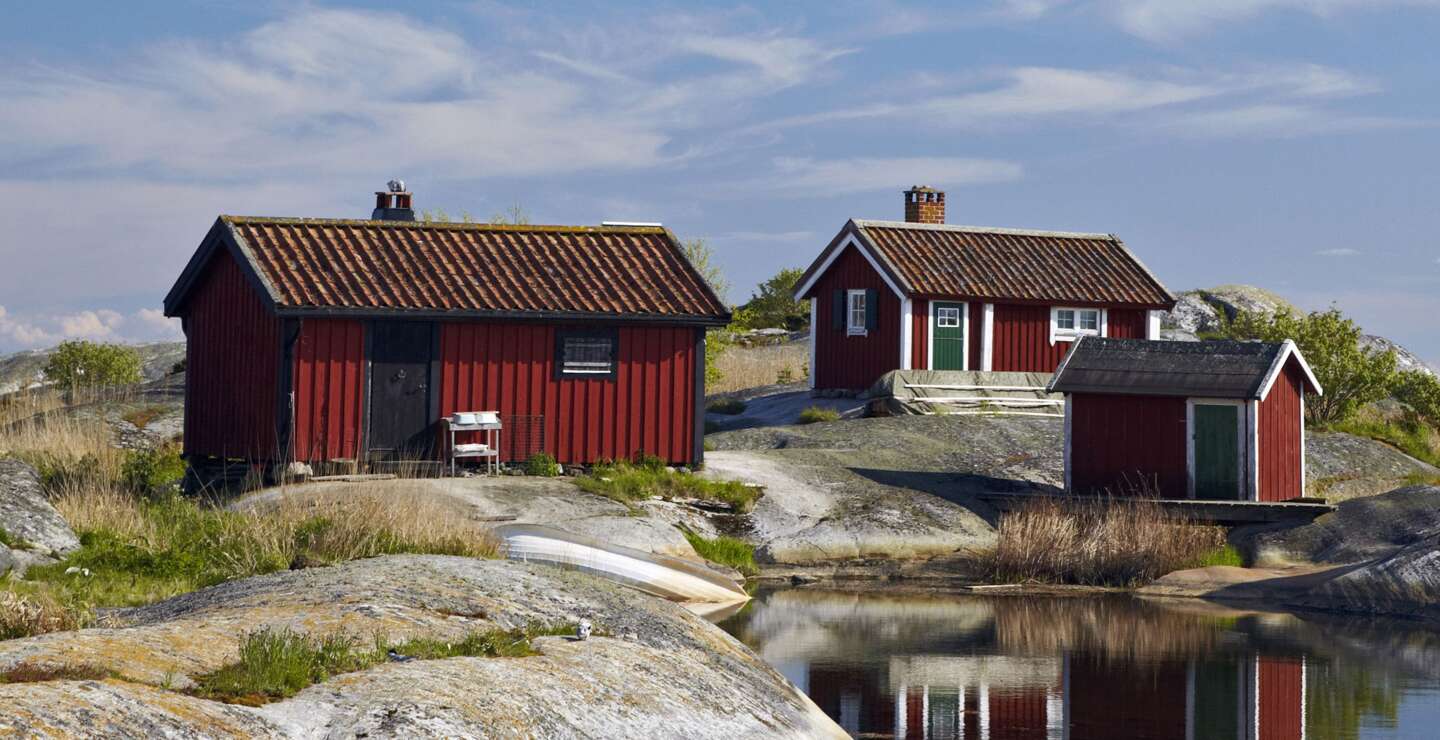 Drei rote Holzhütten mit weißen Fensterrahmen stehen auf Felsen am Wasser. | © imagebank.sweden.se/Ola Ericson