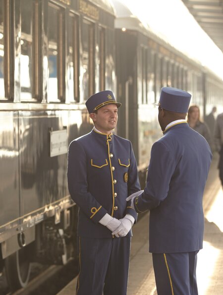 Stewards stehen auf dem Bahnsteig vor dem Venice Simplon Orient Express. Passagiere laufen vorbei. | ©  Matt Hind