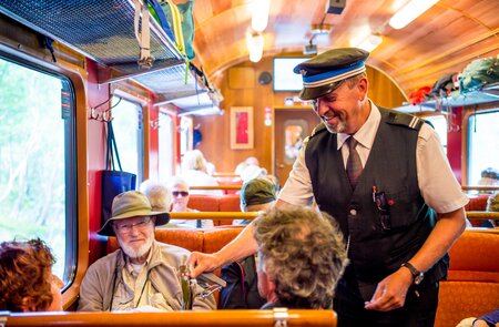 Innenraum eines Zugwagens, in dem Fahrgaeste sitzen und der Zugbegleiter mit einem von ihnen kommuniziert | © Sverre Hjornevik